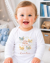 A smiling baby wearing a white onesie with a design of airplanes and the text "Little Captain on board" is sitting in a room with bookshelves and a teddy bear.