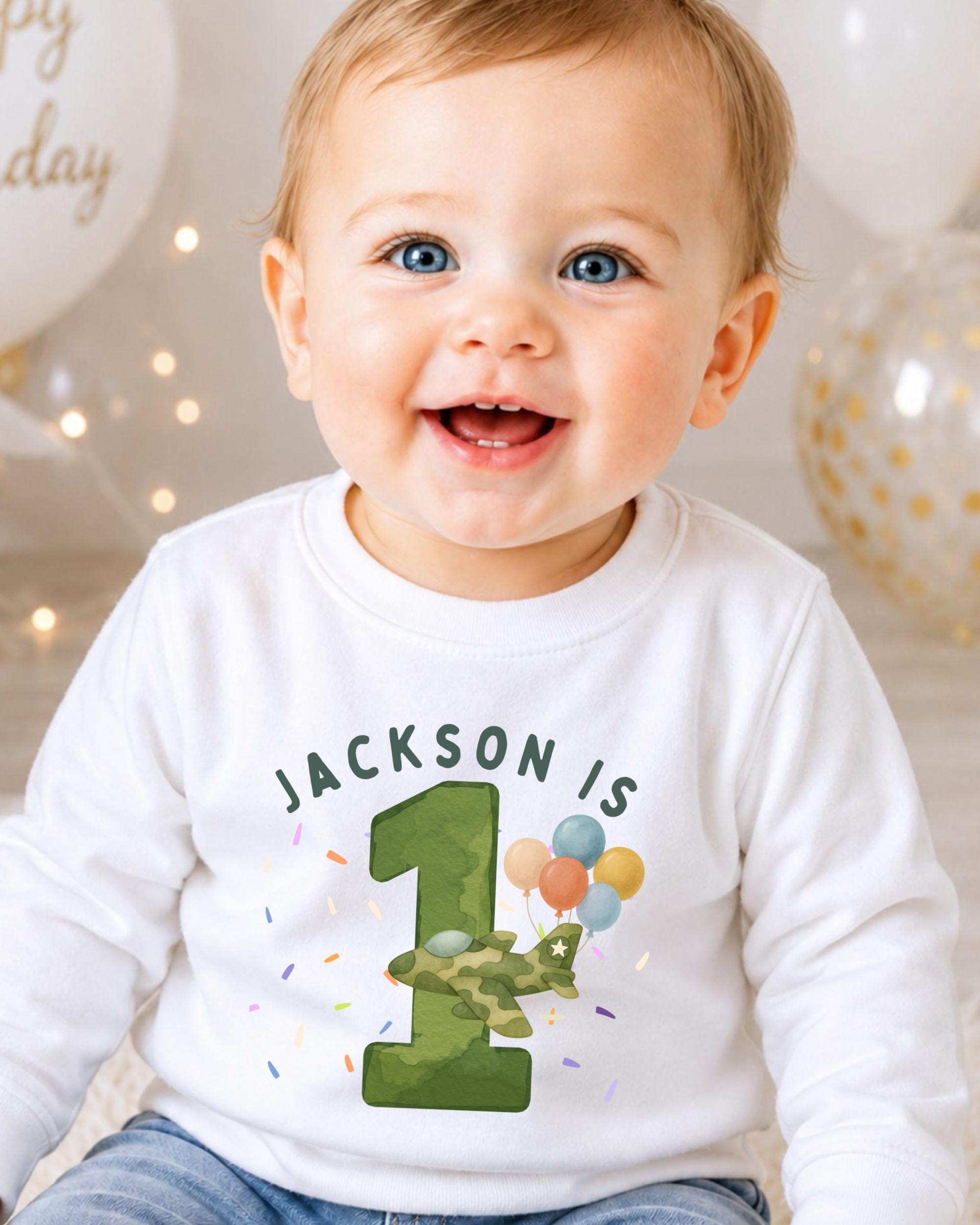 A smiling baby boy wearing a white shirt with a green number one and colorful balloons, celebrating his first birthday.