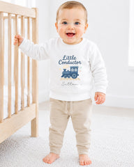 A smiling baby boy wearing a white sweatshirt with a train graphic and the text "Little Conductor" is standing next to a wooden crib.