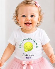 A smiling baby girl wearing a white t-shirt with a tennis ball graphic and the text "First Tennis Match Jessica 2026" printed on it.