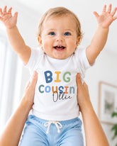 A smiling baby wearing a white t-shirt with the text "BIG COUSIN OLLIE" is being held up by someone's arms.
