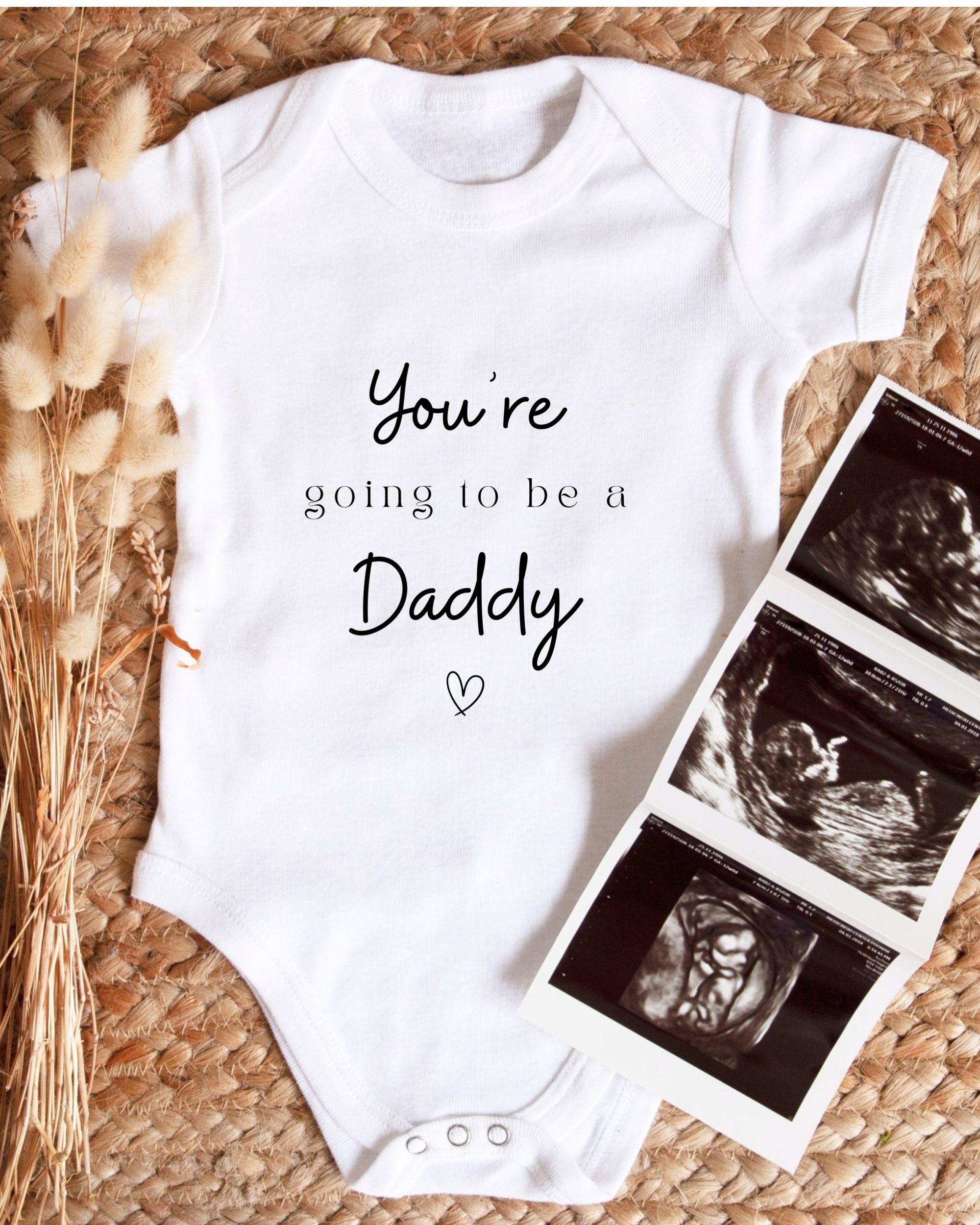 A white baby onesie with the text "You're going to be a Daddy" printed on it, along with a heart symbol. The onesie is placed next to three black and white images of an Ultrasound of a baby's head.