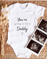 A white baby onesie with the text "You're going to be a Daddy" printed on it, along with a heart symbol. The onesie is placed next to three black and white images of an Ultrasound of a baby's head.