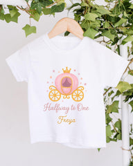 A white t-shirt with a pink and gold illustration of a princess carriage and the text "Halfway to One Freya" printed on it, hanging on a wooden hanger.