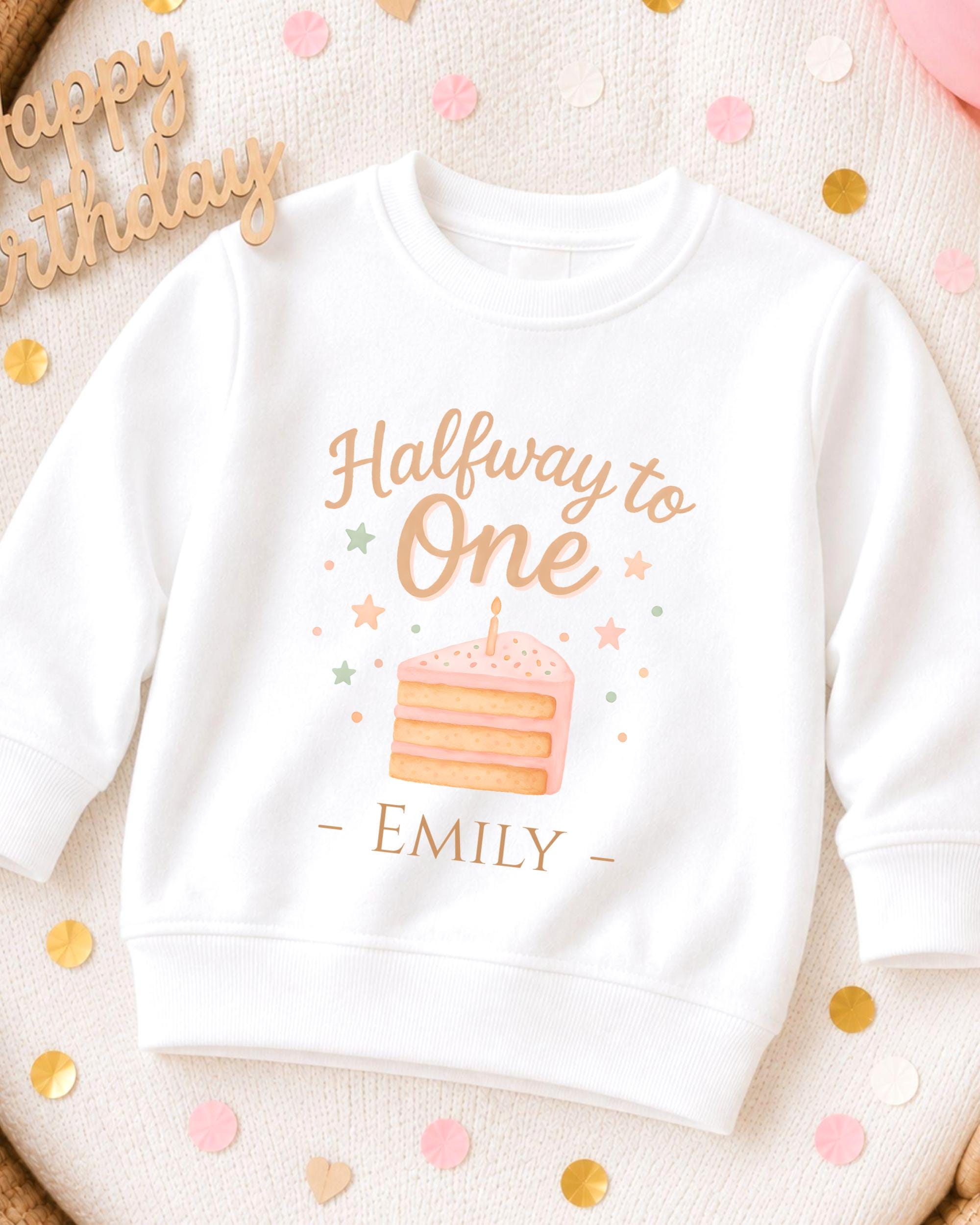 A white sweatshirt with a birthday cake design and the text "Halfway to One" printed on it, along with the name "EMILY" below the cake.