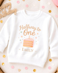 A white sweatshirt with a birthday cake design and the text "Halfway to One" printed on it, along with the name "EMILY" below the cake.