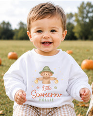 A smiling baby boy wearing a white sweatshirt with a scarecrow graphic and the text "Little Scarecrow Hollie" is sitting in a field with pumpkins in the background.