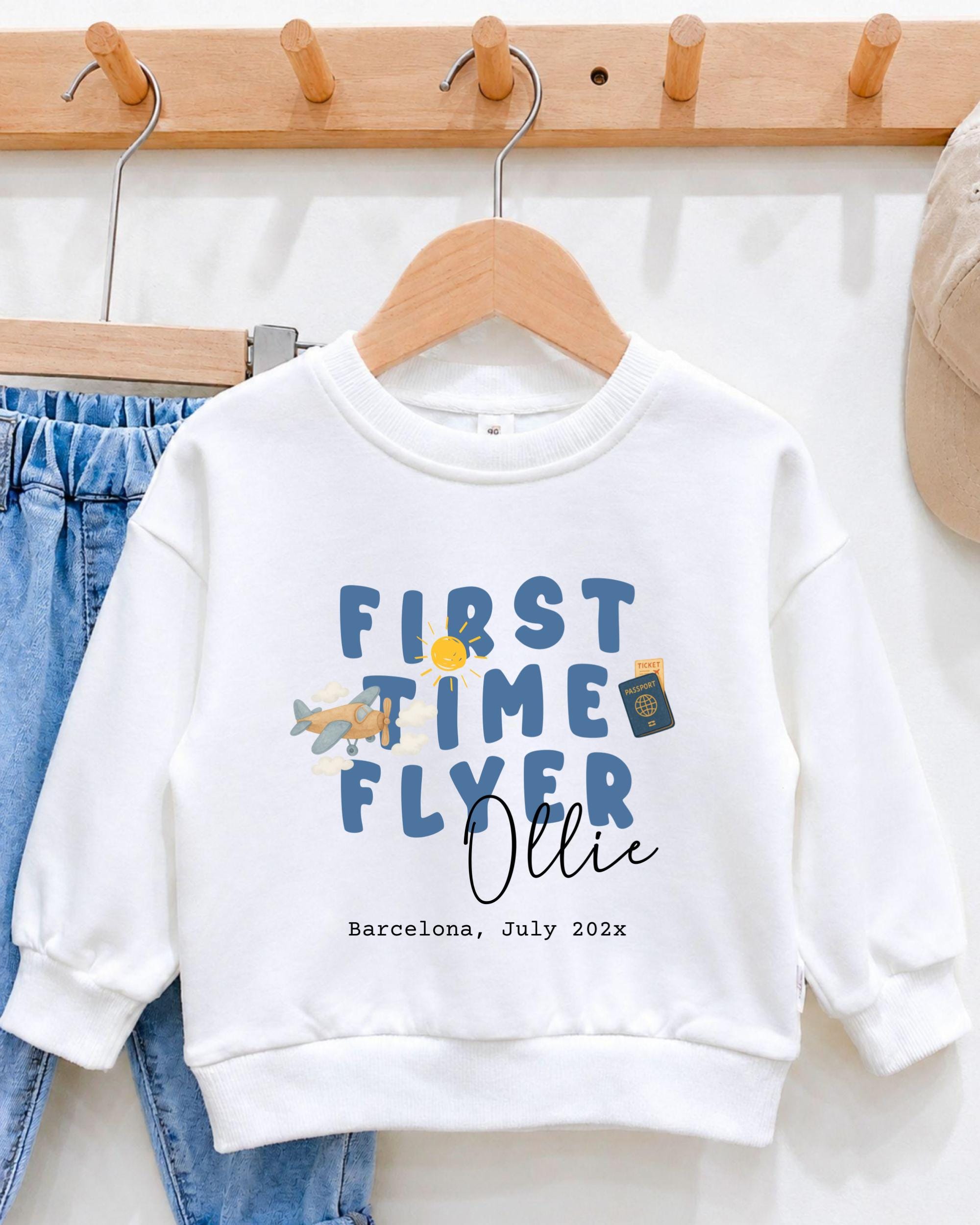 A white sweatshirt with the text "FIRST TIME FLYER OLLIE" printed on it, along with a small airplane and a passport. The sweatshirt is hanging on a wooden hanger, and there is a pair of blue jeans visible in the background.