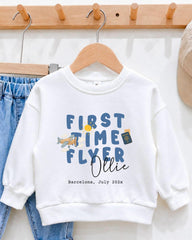 A white sweatshirt with the text "FIRST TIME FLYER OLLIE" printed on it, along with a small airplane and a passport. The sweatshirt is hanging on a wooden hanger, and there is a pair of blue jeans visible in the background.