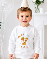 A young boy wearing a white sweatshirt with a giraffe and balloons on it, smiling at the camera.