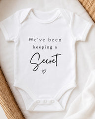 A white baby onesie with the text "We've been keeping a Secret" printed on it.