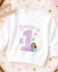 A white sweatshirt with a mermaid and a number 1 printed on it, surrounded by confetti.