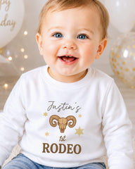A smiling baby boy wearing a white shirt with a graphic design and text.
