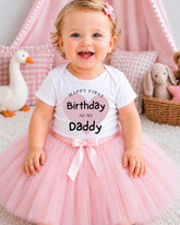 A smiling baby girl wearing a white t-shirt with text and a pink tutu, sitting on a pink carpet with stuffed animals in the background.