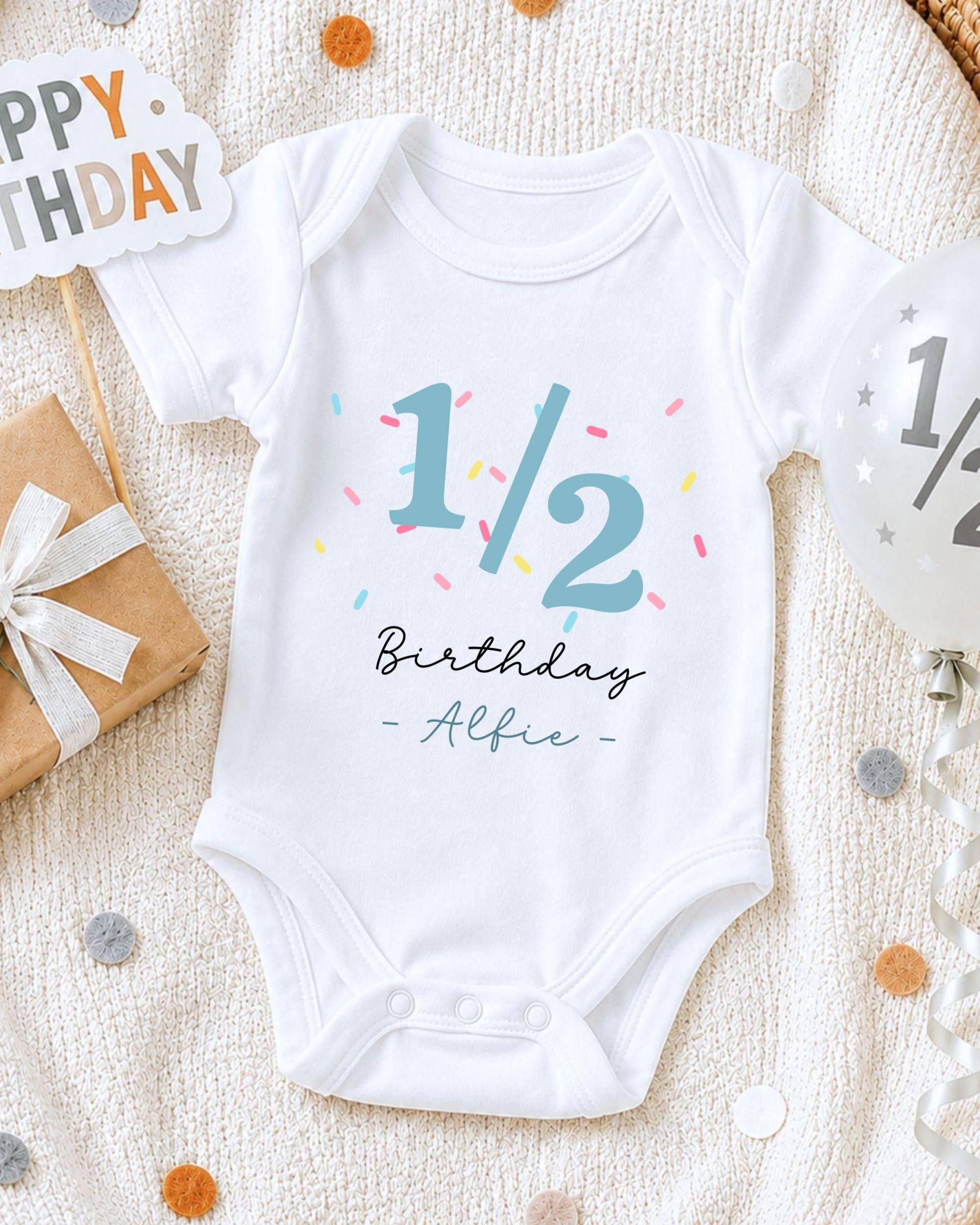 A white onesie with the text "1/2 Birthday - Alfie" printed on it, along with a birthday sign and confetti, is displayed on a bed.