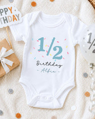 A white onesie with the text "1/2 Birthday - Alfie" printed on it, along with a birthday sign and confetti, is displayed on a bed.