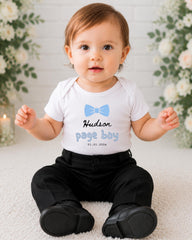 A smiling baby boy wearing a white shirt with a blue bow tie and black pants sits on a white carpet, with a floral background behind him.