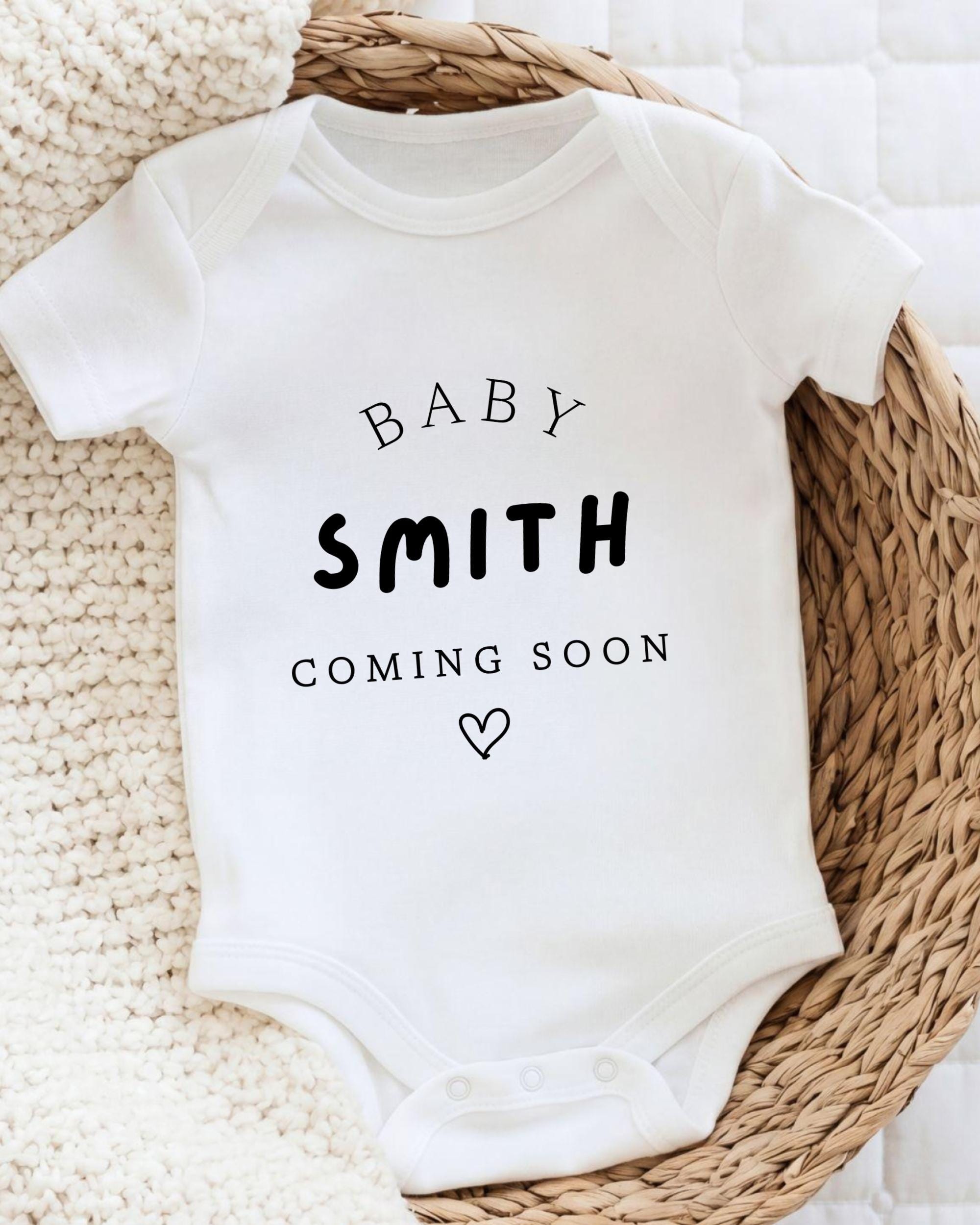 A white baby onesie with the text "BABY SMITH COMING SOON" printed on it, along with a heart symbol.