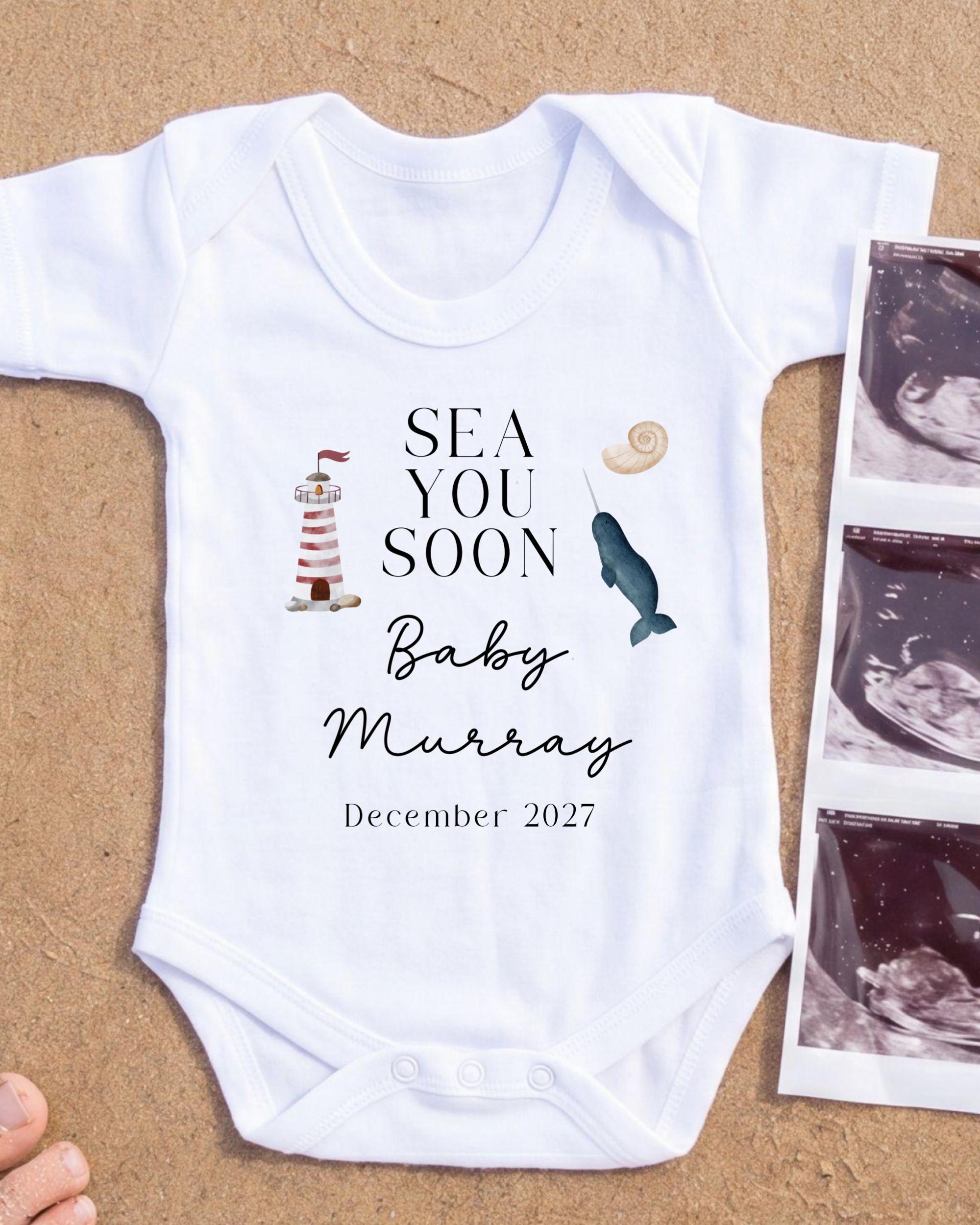A white onesie with text and illustrations related to a baby announcement, placed on a sandy surface next to a photograph of an Ultrasound image.