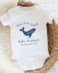 A white baby onesie with a blue whale graphic and text that reads "Sea You Soon Baby Seashell Due December 2021".