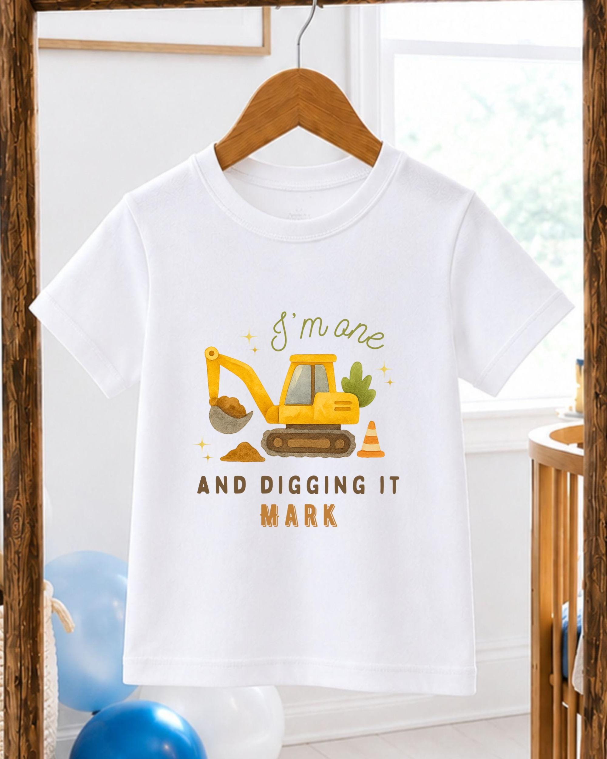 A white t-shirt with a graphic design of a yellow construction vehicle and the text "I'm one AND digging it MARK" printed on it.