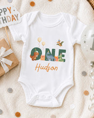 A white onesie with the name "Hudson" printed on it, surrounded by various birthday decorations such as balloons, a gift box, and confetti.
