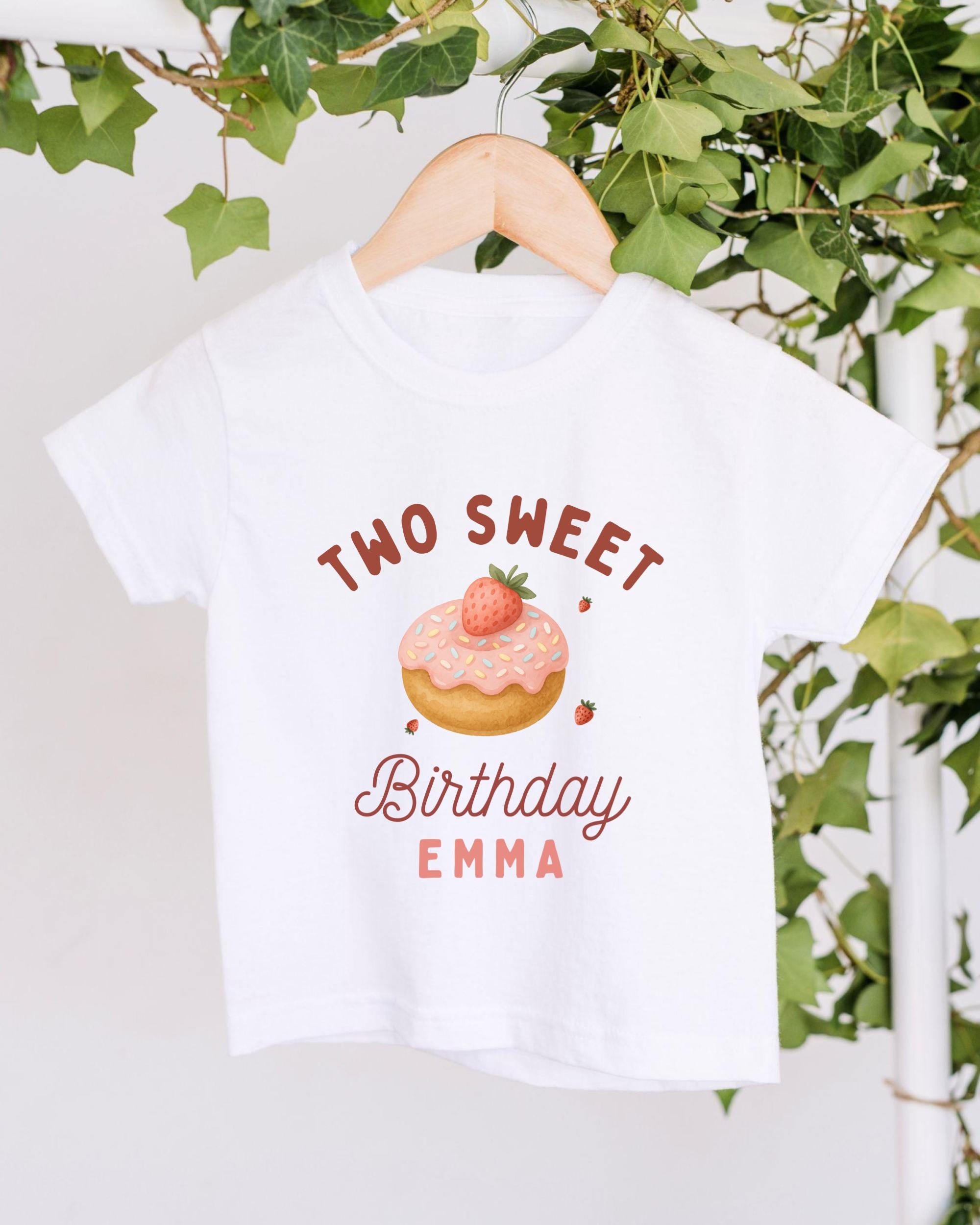 A white t-shirt with a graphic design featuring a strawberry donut and the text "Two Sweet Birthday Emma" printed on it.