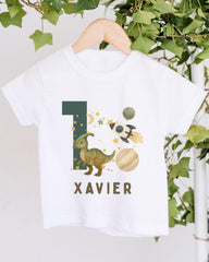 A white t-shirt with a dinosaur graphic and the number 1, along with a rocket ship and stars, hanging on a wooden hanger.