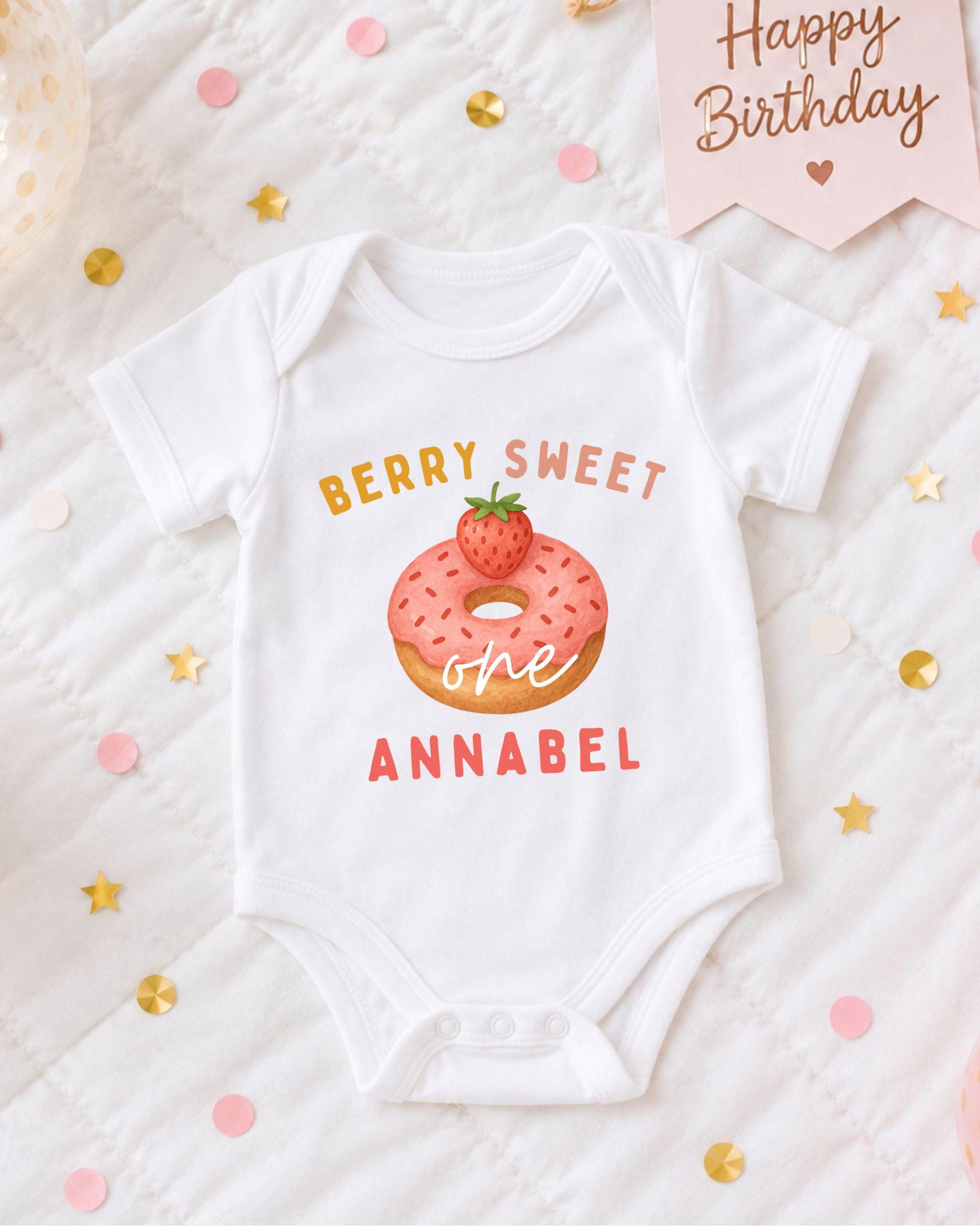 A white onesie with a pink donut and the text "Berry Sweet one Annabel" printed on it, surrounded by confetti and a birthday card.