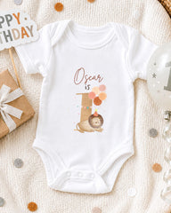A white onesie with the text "Oscar is" printed on it, along with a lion and balloons, is displayed on a bed with a "Happy Birthday" sign and a birthday balloon.