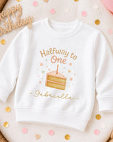 A white sweatshirt with a birthday cake design and the text "Halfway to One Gabriella" printed on it, surrounded by confetti.
