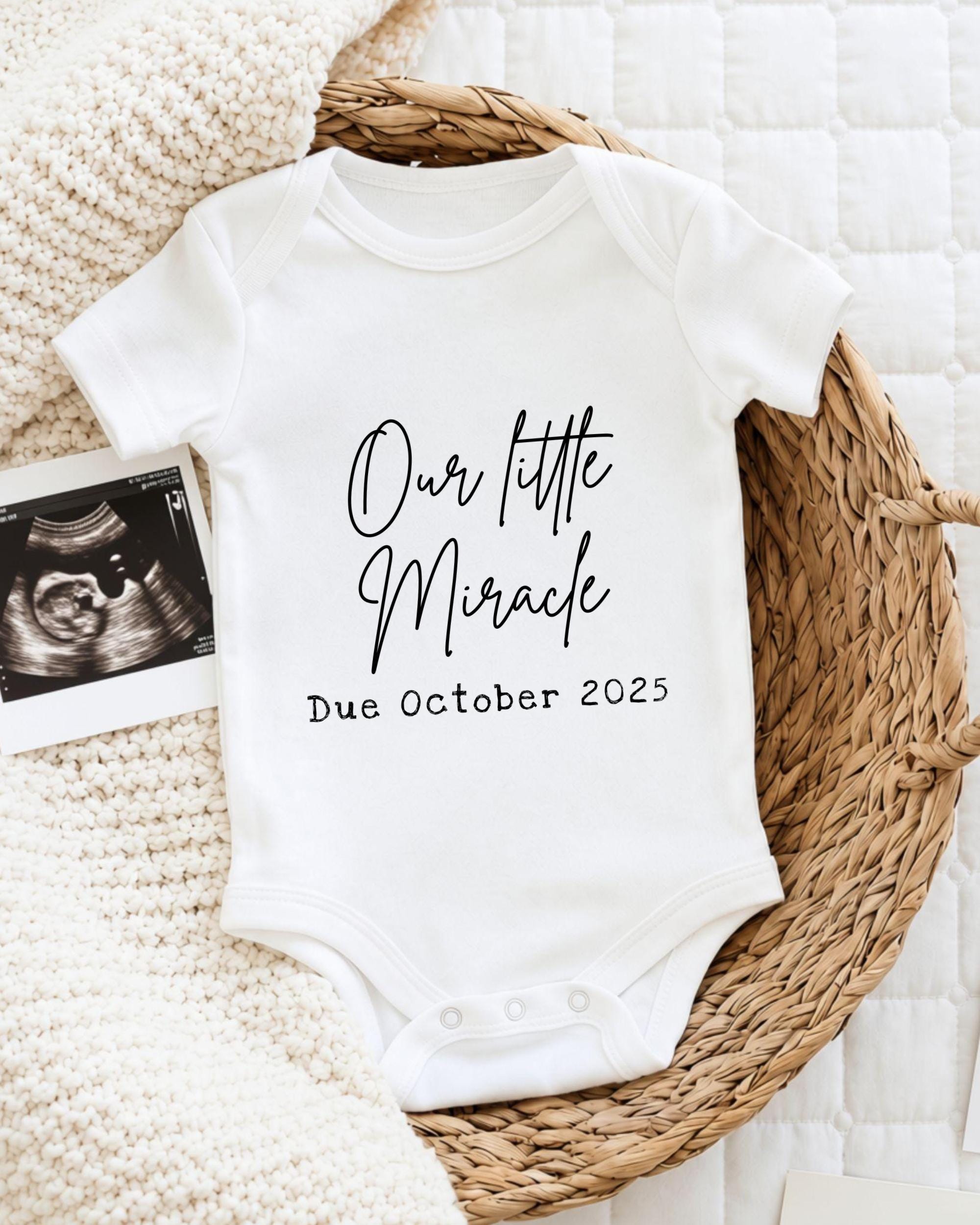 A white baby onesie with the text "Our little Miracle Due October 2025" printed on it, placed on a woven basket.