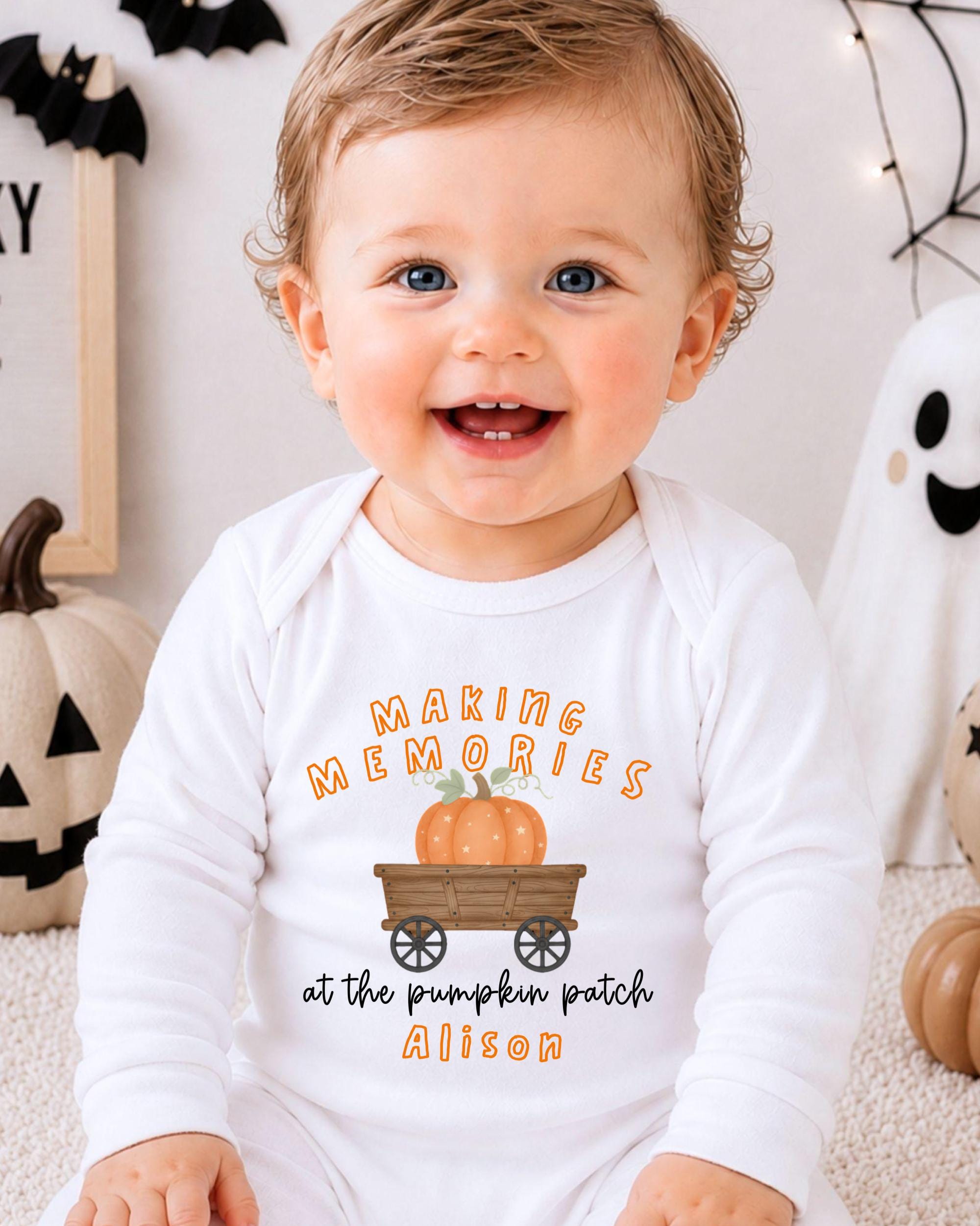 A smiling baby wearing a white onesie with a pumpkin and carriage graphic, sitting in front of Halloween decorations.