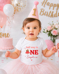 A young girl wearing a birthday hat and a white t-shirt with the text "half way to one" and "Sophie" is posing in front of a birthday cake and balloons.