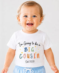 A smiling baby wearing a white t-shirt with text that says "I'm Going to be a Big Cousin Est 2029".