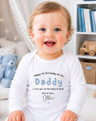 A smiling baby boy wearing a white onesie with a personalized message, sitting in a room with a teddy bear and bookshelf in the background.