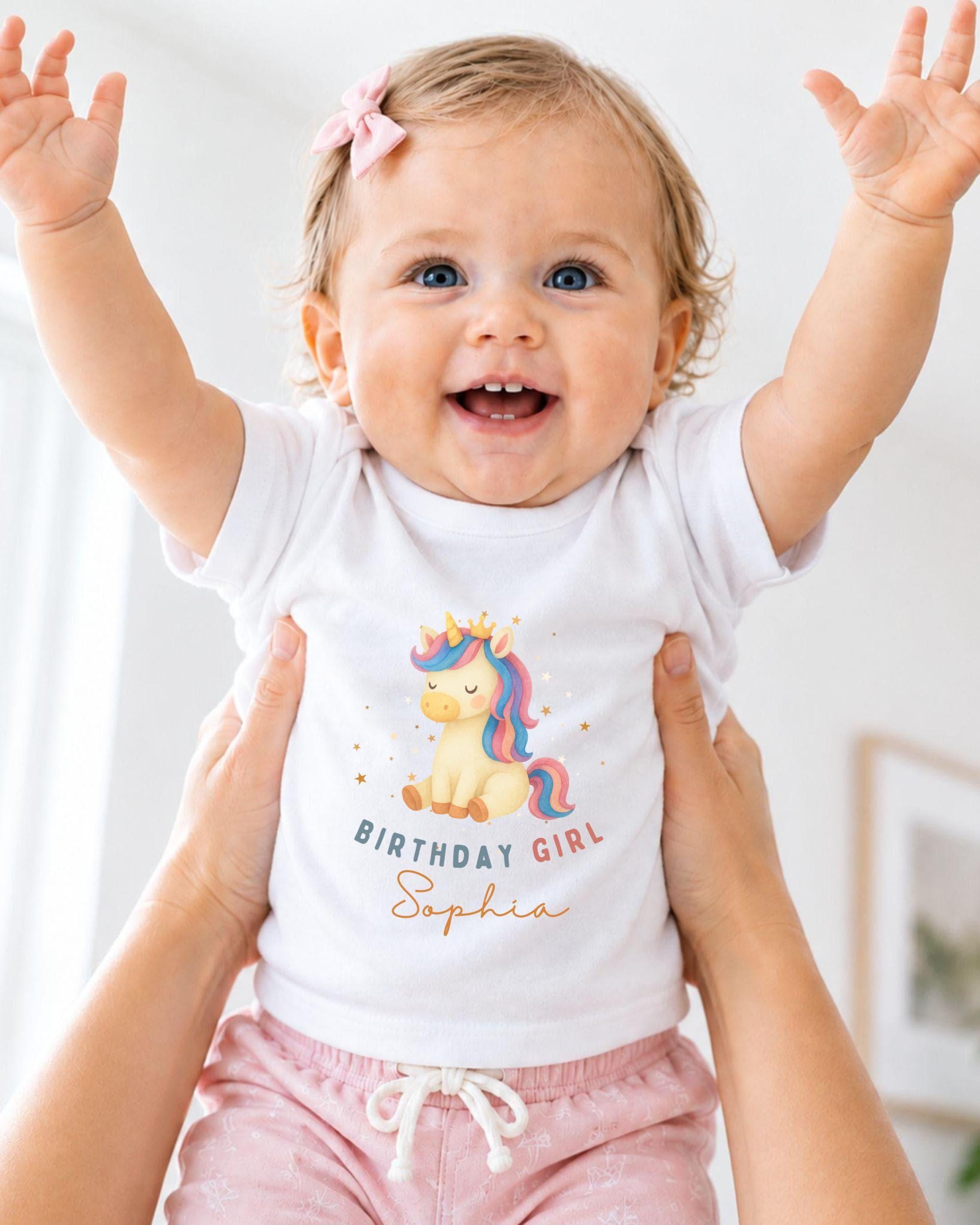 A smiling baby girl wearing a white t-shirt with a unicorn graphic and the text "Birthday Girl Sophia" is being held up by someone's arms.