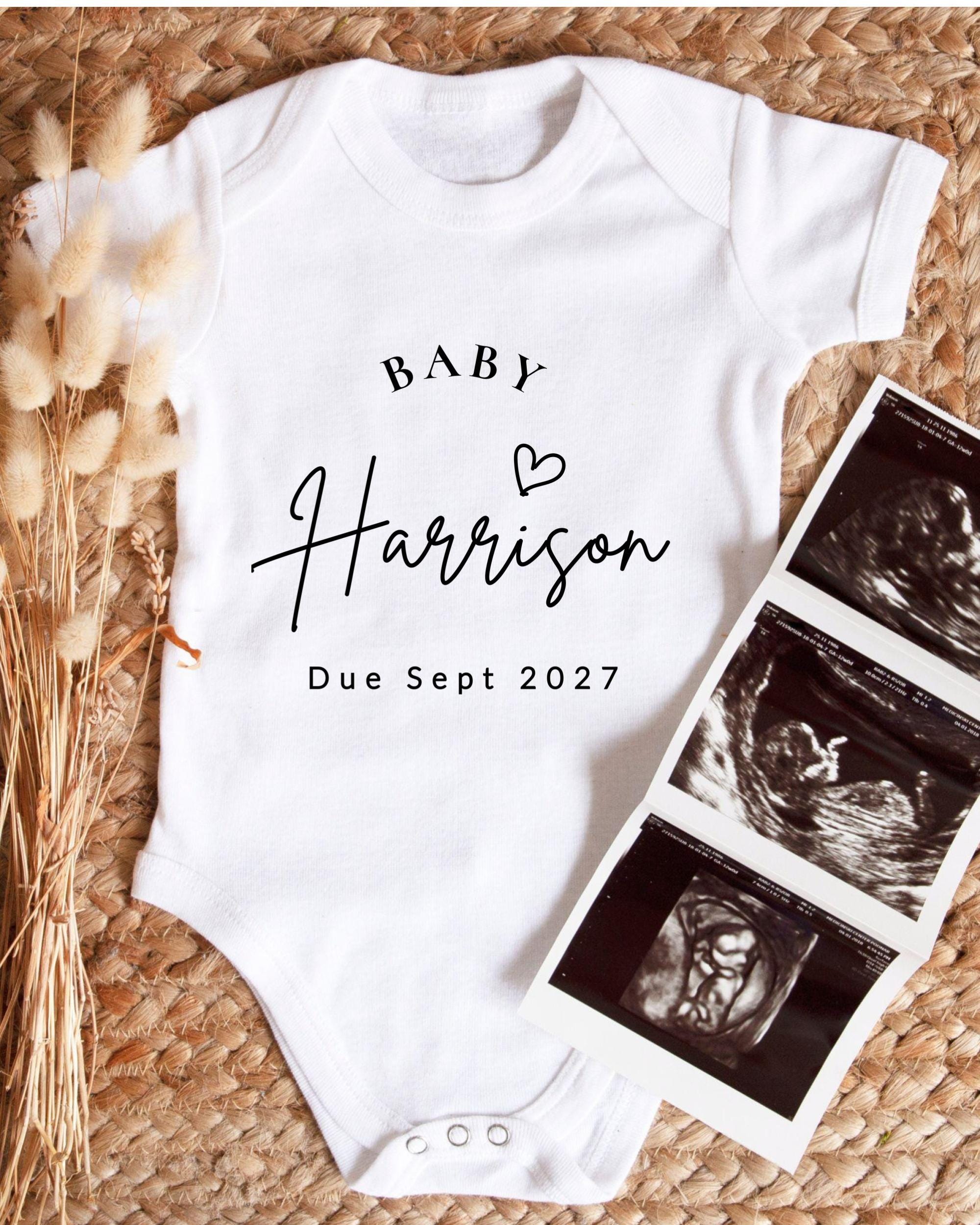 A white baby onesie with the text "BABY Harrison Due Sept 2027" printed on it, along with a photo of an ultrasound image.