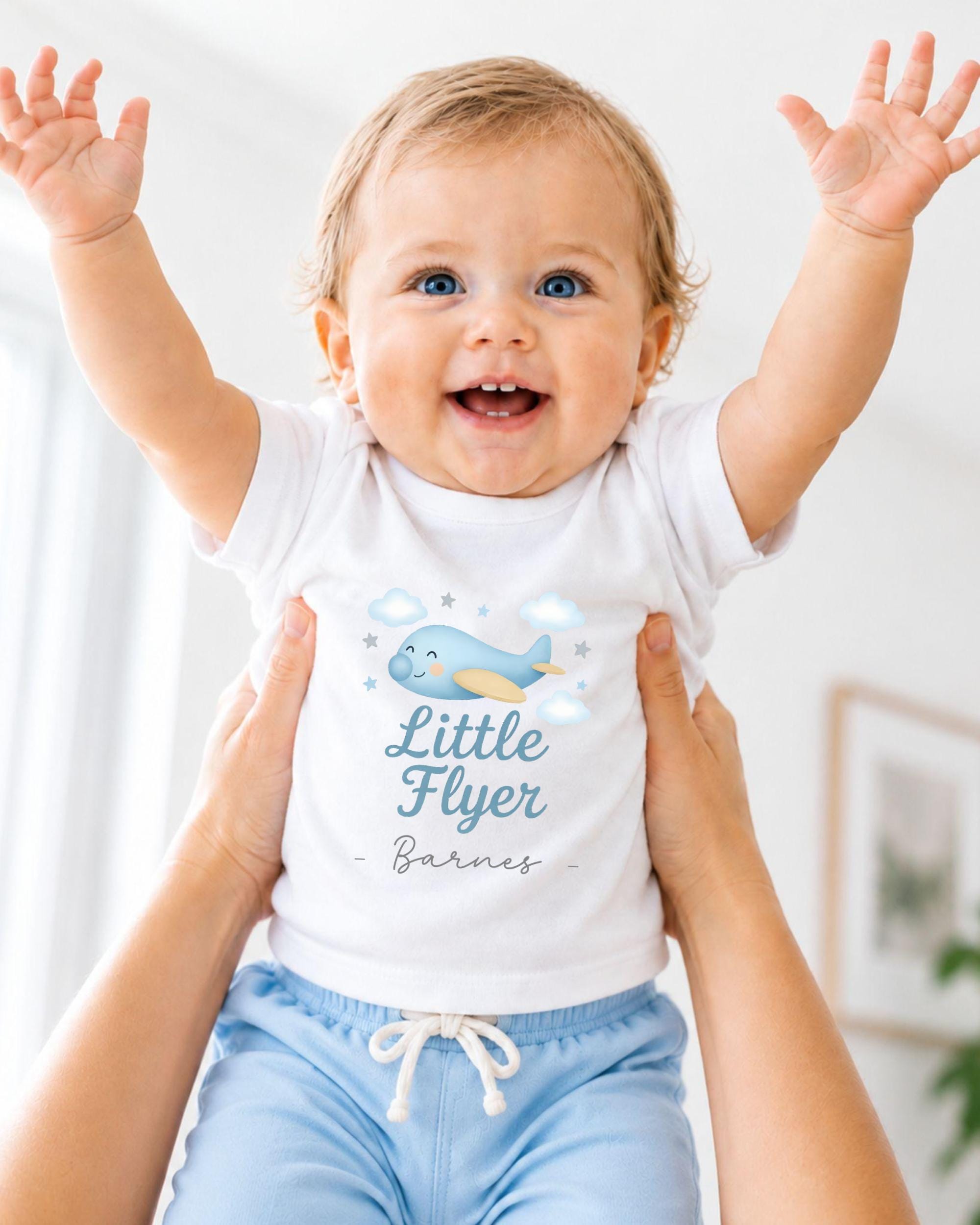 A smiling baby wearing a white shirt with a cartoon airplane and the text "Little Flyer" is being held up by someone's arms.