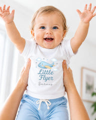 A smiling baby wearing a white shirt with a cartoon airplane and the text "Little Flyer" is being held up by someone's arms.