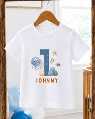 A white t-shirt with a graphic design featuring a blue jellyfish, a starfish, and other sea creatures, along with the text "JOHNNY" printed on it.