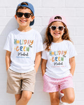Two children wearing matching white t-shirts with text and graphics, smiling and posing against a white brick wall.