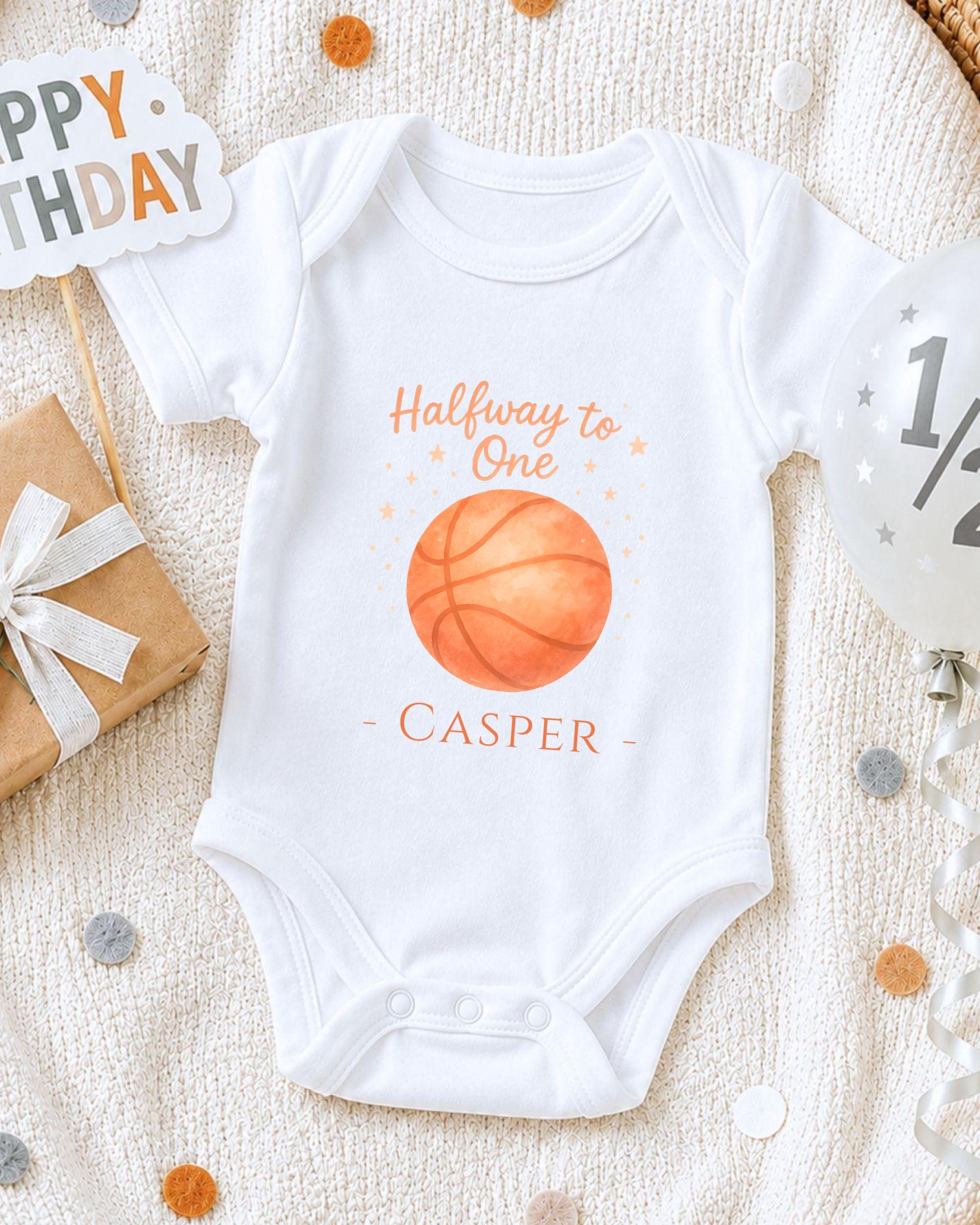 A white onesie with a basketball design and the text "Halfway to One - CASPER" printed on it, along with a "Happy Birthday" sign and a balloon.