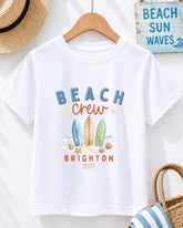 A white t-shirt with a graphic design featuring three surfboards and beach elements, along with the text "BEACH CREW BRIGHTON 2027".