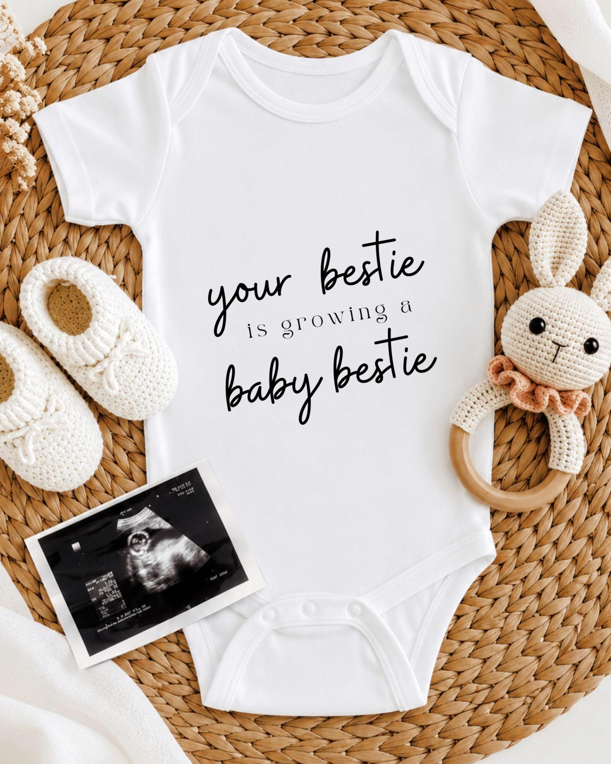 A white onesie with the text "your bestie is growing a baby bestie" printed on it, along with a crocheted bunny toy and a baby shoe. There is also an Ultrasound image of a baby's belly.