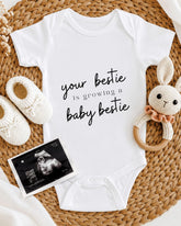 A white onesie with the text "your bestie is growing a baby bestie" printed on it, along with a crocheted bunny toy and a baby shoe. There is also an Ultrasound image of a baby's belly.