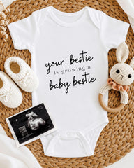 A white onesie with the text "your bestie is growing a baby bestie" printed on it, along with a crocheted bunny toy and a baby shoe. There is also an Ultrasound image of a baby's belly.