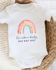 A white baby onesie with a colorful rainbow graphic and the text "Rainbow baby due novely 2027" printed on it.