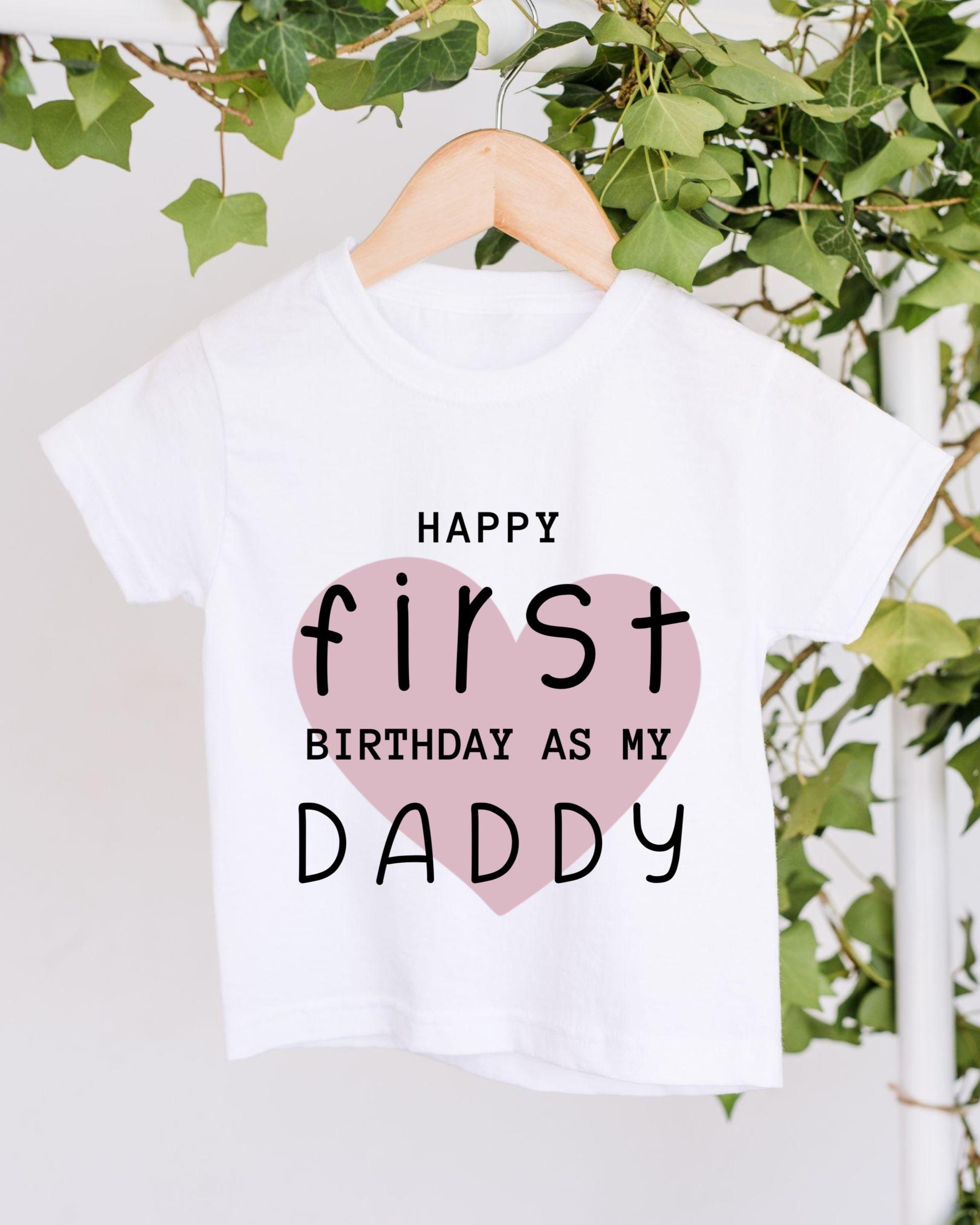 A white t-shirt with a pink heart and text that reads "HAPPY FIRST BIRTHDAY AS MY DADDY" hanging on a wooden hanger.