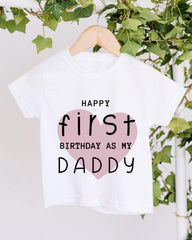 A white t-shirt with a pink heart and text that reads "HAPPY FIRST BIRTHDAY AS MY DADDY" hanging on a wooden hanger.